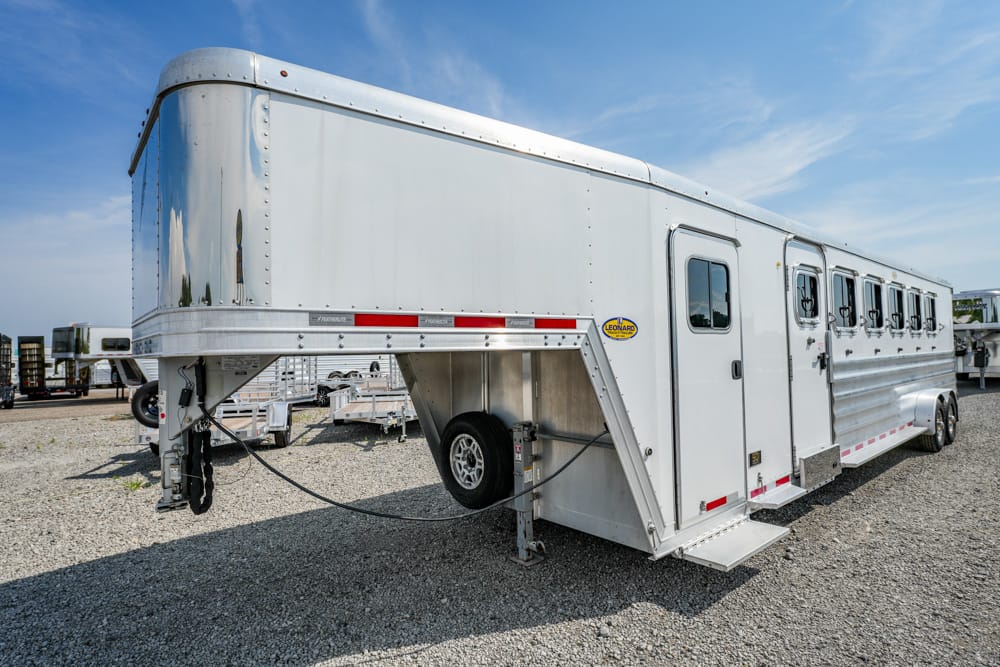 White aluminum gooseneck horse trailer parked on a gravel lot, with side doors, windows, and a spare tire mounted under the front section
