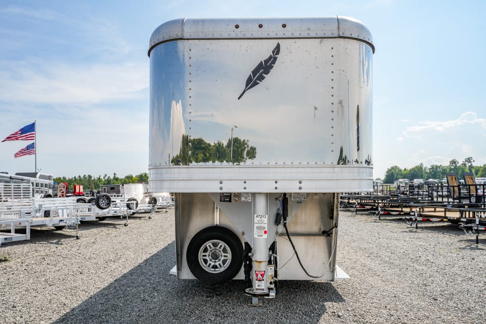 Front view of a polished aluminum trailer with a feather logo, parked in a trailer lot with other trailers and American flags in the background.