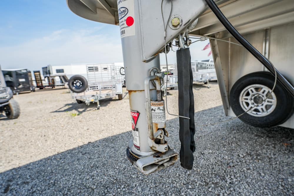 Close-up of a travel trailer jack with a black strap, rust marks, and warning stickers on a gravel lot.
