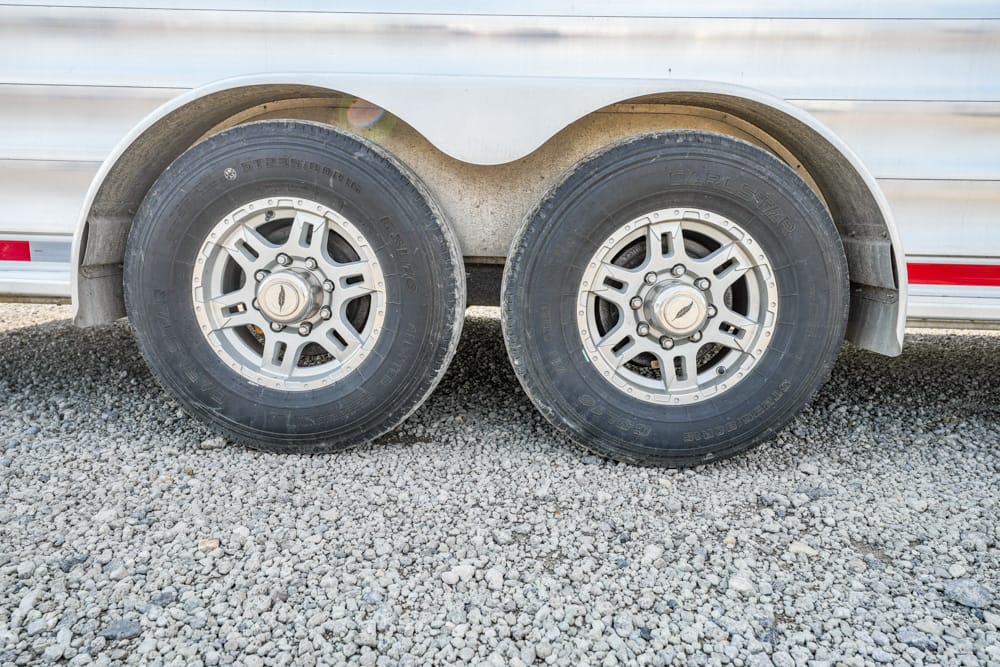 Dual trailer wheels with silver rims on a gravel surface under a silver trailer.