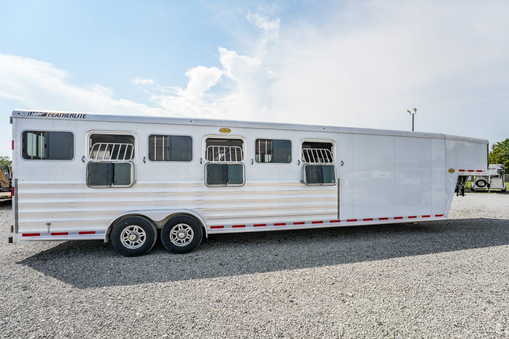 Long white horse trailer with multiple windows and dual axles, parked on a gravel lot under a blue sky with scattered clouds.