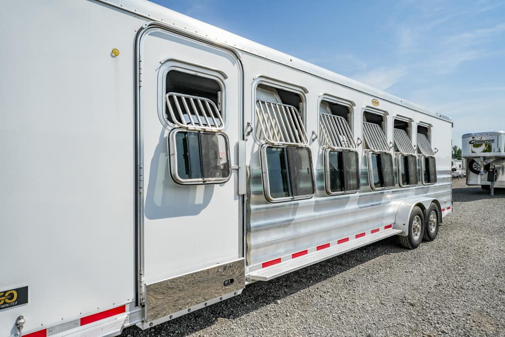 White horse trailer with ventilated side windows parked on a gravel lot; another trailer is in the background.
