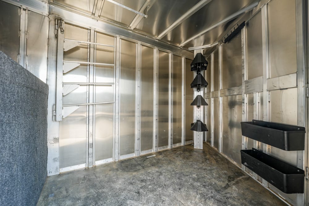 Empty metal horse trailer interior with wall-mounted saddle racks at the back and two black storage bins on the right wall