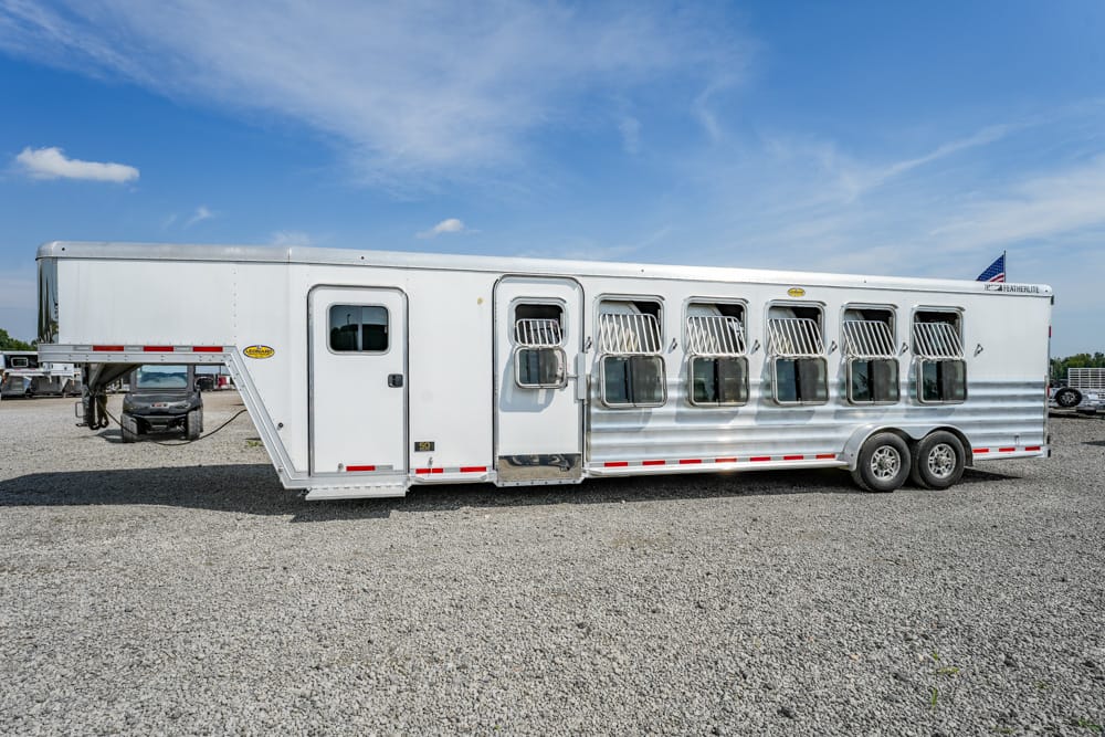 Long aluminum horse trailer with seven side windows and a front access door, parked on a gravel lot beneath a blue sky.