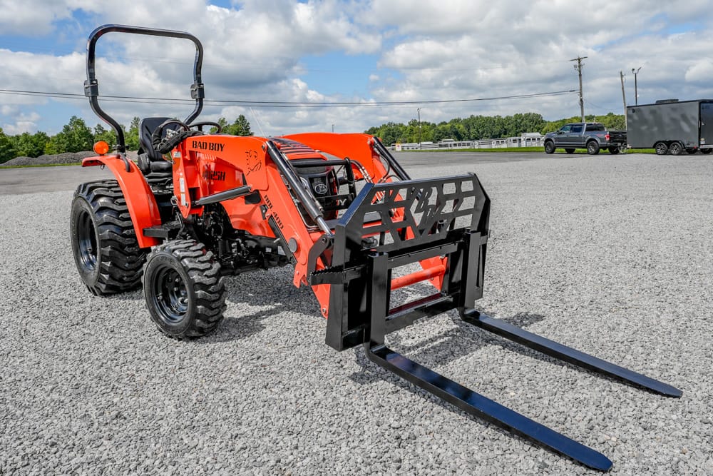 Orange compact tractor with a black front loader and fork attachment parked on a gravel lot.