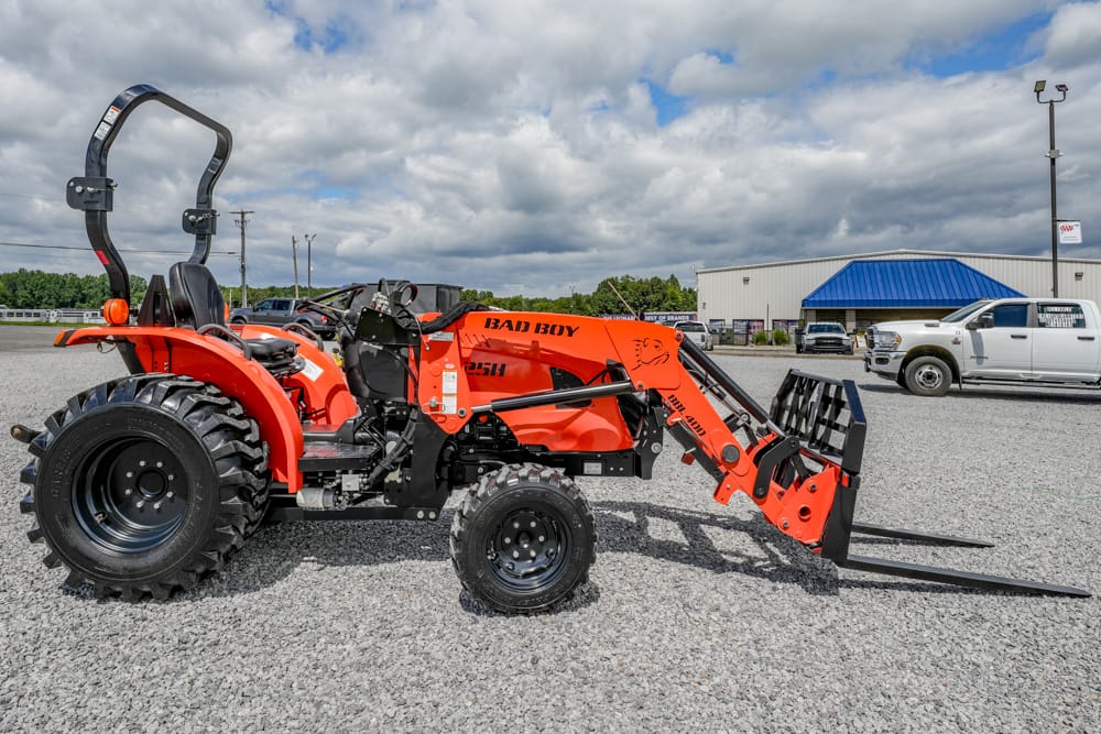 Orange Bad Boy compact tractor with a front loader and forks on a gravel lot, large rear tires and roll bar visible.