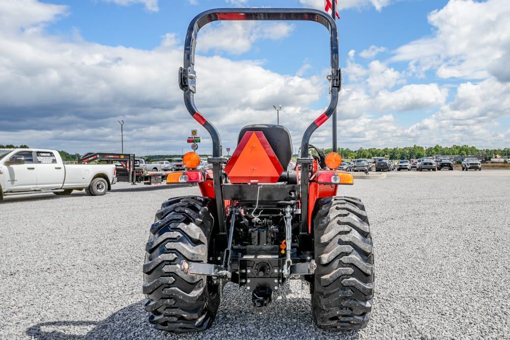 Rear view of a red tractor in a gravel lot, a triangular slow‑moving vehicle sign mounted on the back, blue sky above with clouds.
