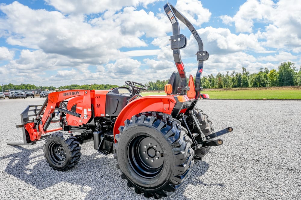 Orange compact utility tractor with front loader attachment parked on gravel, ROPS bar and large rear tires visible in a field setting