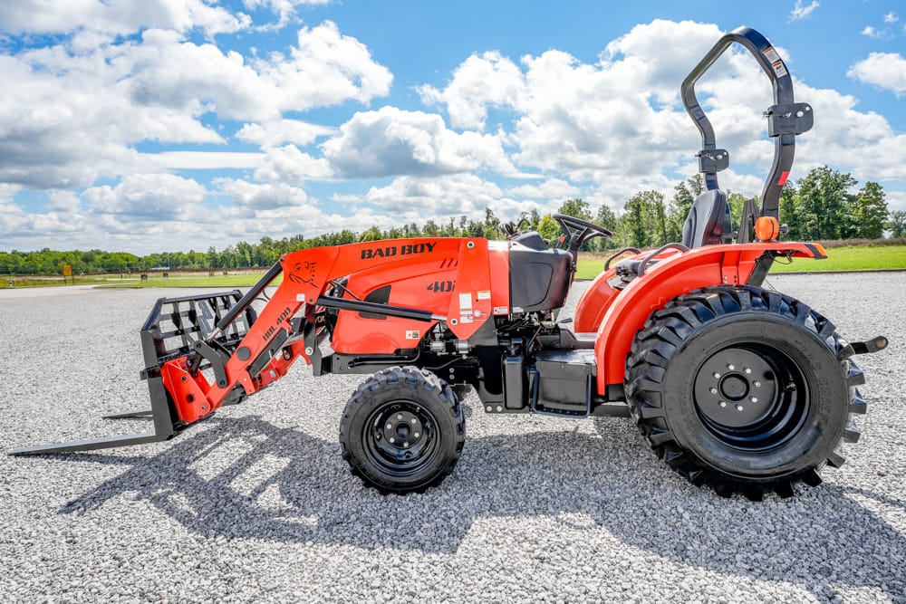 Orange Bad Boy utility tractor with front loader and pallet forks on a gravel surface under a blue sky with clouds, side view.