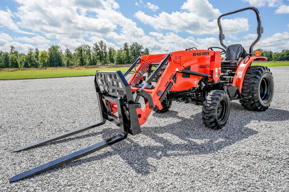 Red Bad Boy utility tractor with a front pallet fork attachment on a gravel lot under a blue, partly cloudy sky.