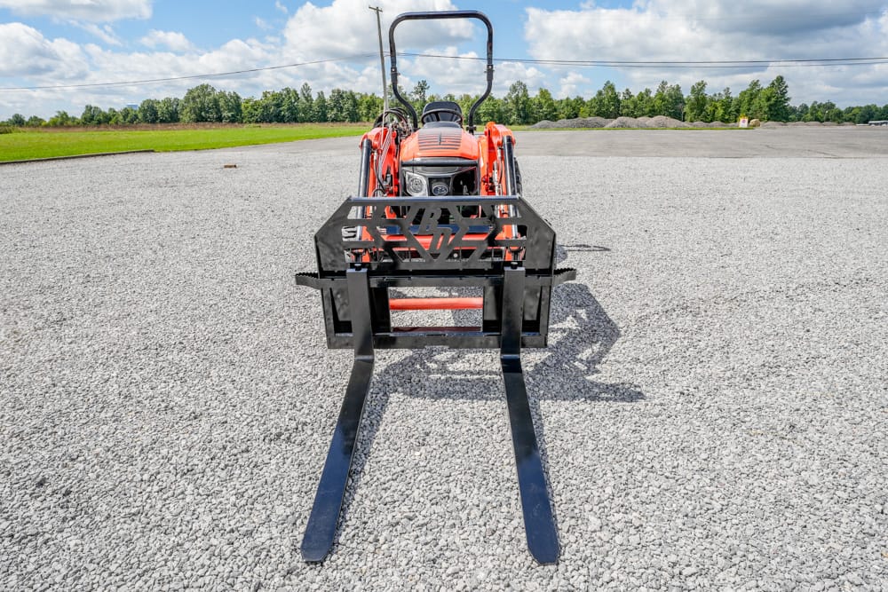 Red tractor with a black pallet-fork attachment on a gravel lot under a partly cloudy blue sky.