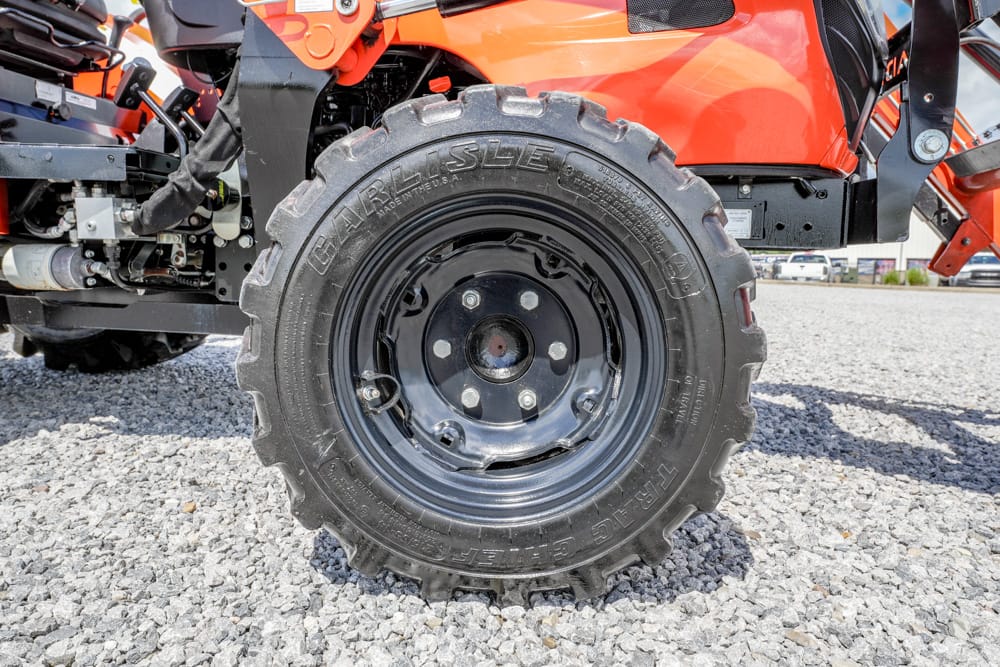 Close-up of a large knobby off-road tire mounted on an orange utility vehicle on a gravel surface, showing the wheel and lug nuts nearby factory components.