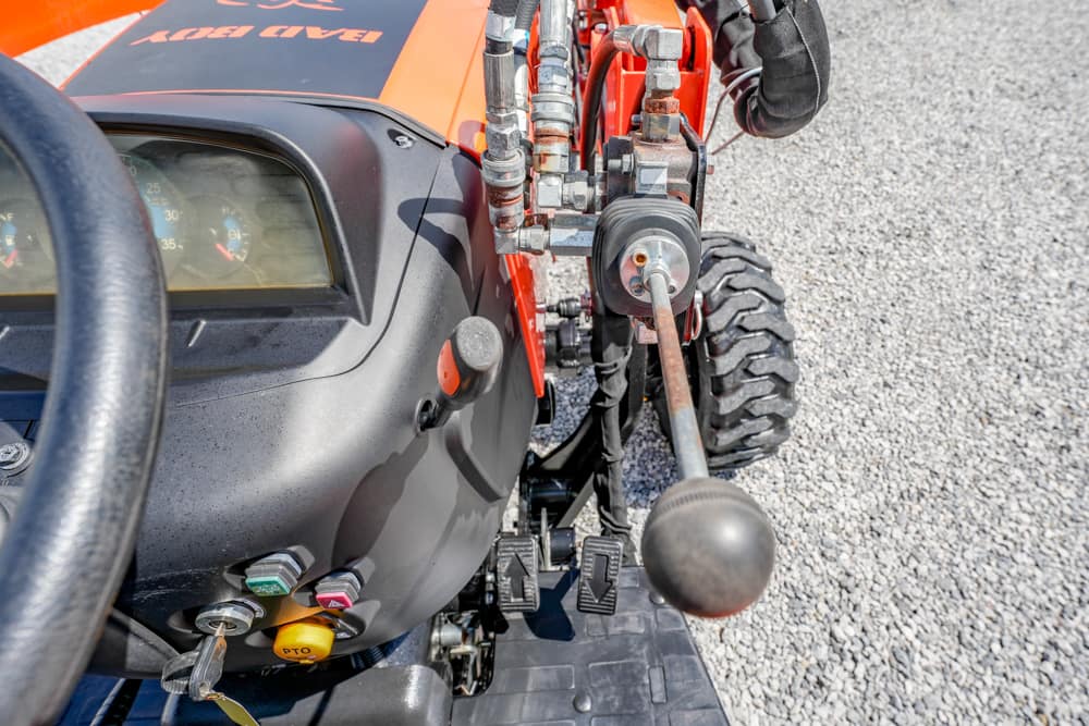 Close-up of a tractor cockpit: steering wheel on the left, dashboard gauges, and a long gear lever with a black knob. nearby colored buttons and controls are visible.