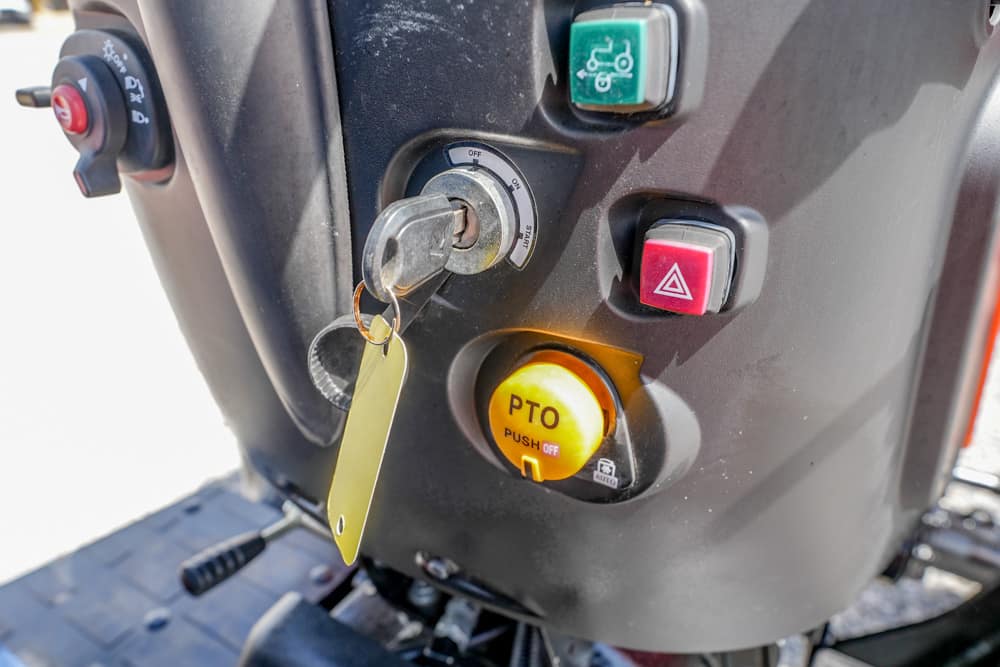 Close-up of a scooter's control panel: ignition with key, yellow PTO push-off button, green maintenance switch, and red hazard button nearby.