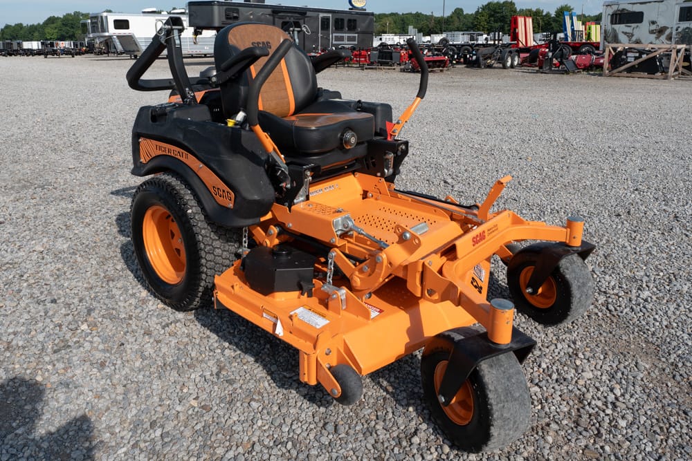 Orange SCAG Tiger Cat commercial zero-turn mower with a black seat on a gravel lot, trailers visible in the background.