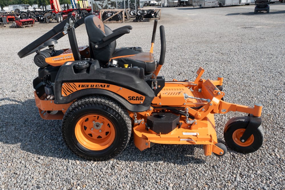 Orange SCAG Tiger Cat II riding mower with a wide front cutting deck, parked on a gravel lot at an equipment yard.