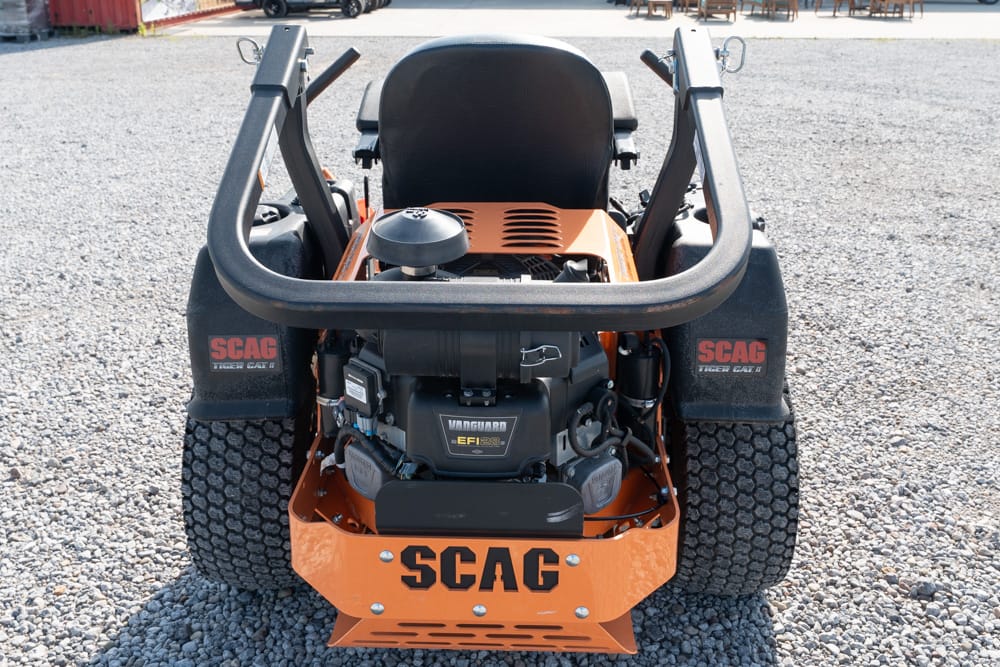 Rear view of an orange SCAG ride-on lawn mower on gravel, with exposed engine and protective roll bar visible.