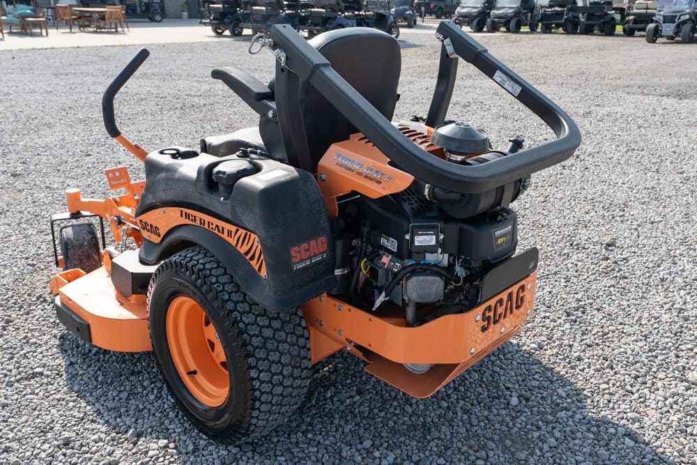 Orange SCAG riding lawn mower on a gravel lot with exposed engine and large tires.
