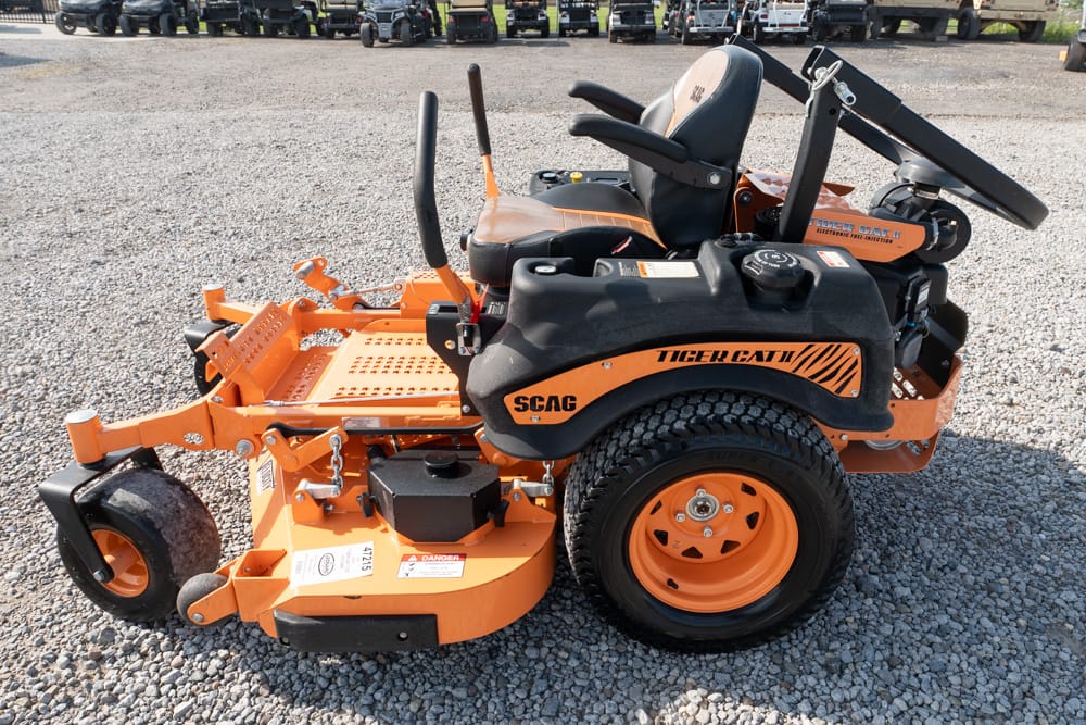 Orange Scag Tiger Cat II riding lawn mower with a large cutting deck parked on a gravel lot on a sunny day.