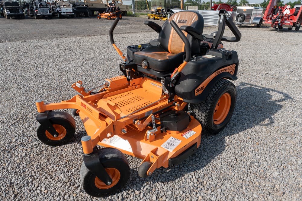 Orange SCAG zero-turn riding lawn mower with a wide cutting deck and black cushioned seat on a gravel lot.