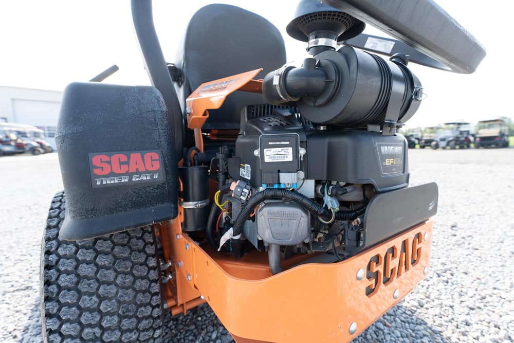 Close-up of the front section of an orange SCAG Tiger Cat II turf maintenance machine, showing the engine, hoses, and a large wheel with a textured tread.