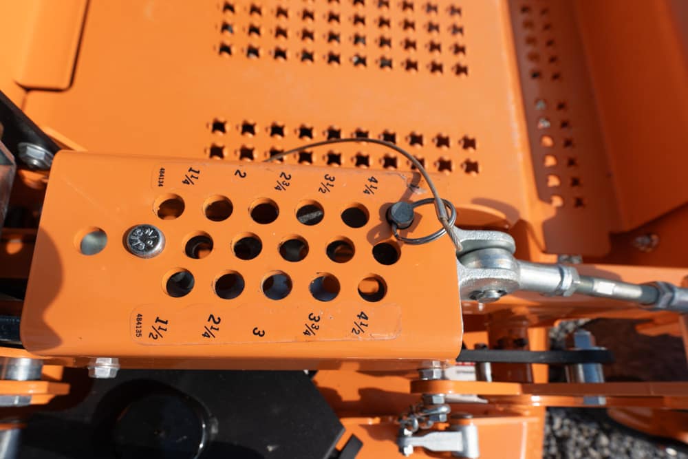 Close-up of orange industrial machinery showing a perforated plate with numbered holes and a hydraulic linkage