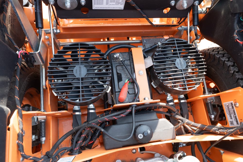 Close-up of an orange industrial machine's engine bay showing two large round cooling fans, wiring, and a battery.
