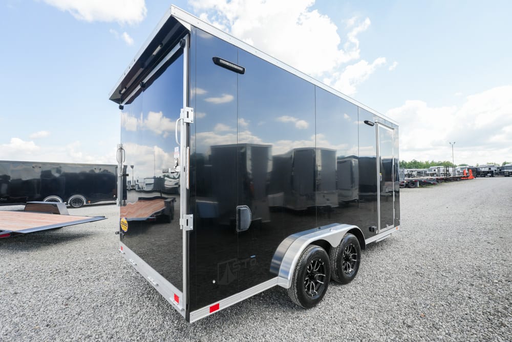 Black tandem-axle enclosed cargo trailer parked on a gravel lot, reflecting a blue sky and clouds in its glossy panels, rear side visible.
