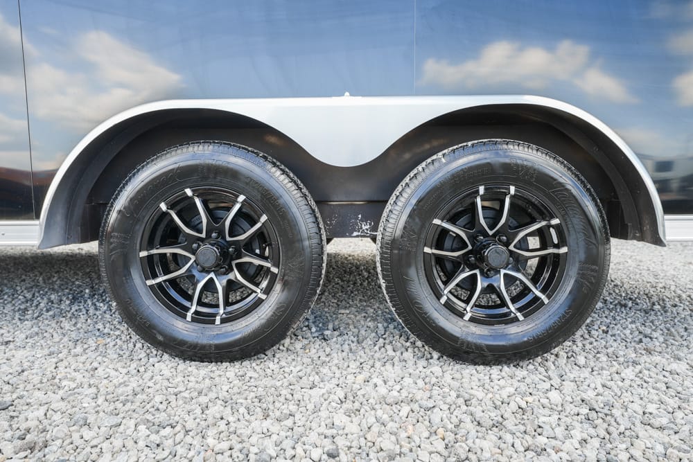 Dual-axle trailer wheels with black rims on a gravel surface under a dark trailer side.