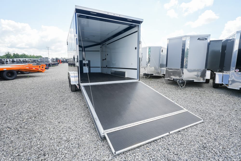 Open cargo trailer with ramp lowered for loading, parked on a gravel lot with other trailers nearby.