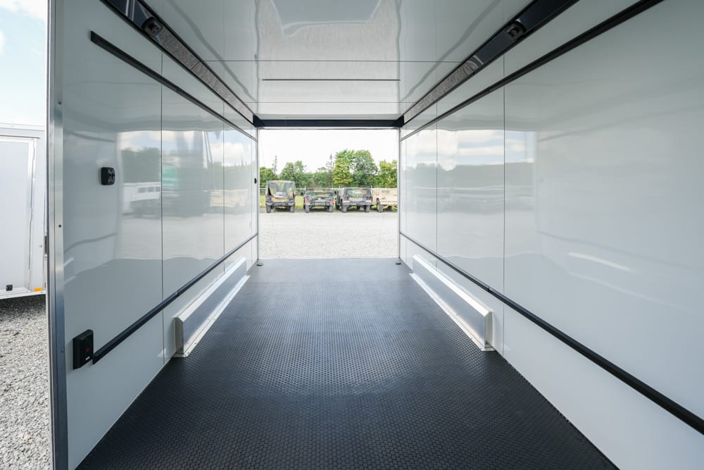 Interior of an empty white cargo trailer, looking toward the open back to a gravel lot with parked vehicles and trees beyond