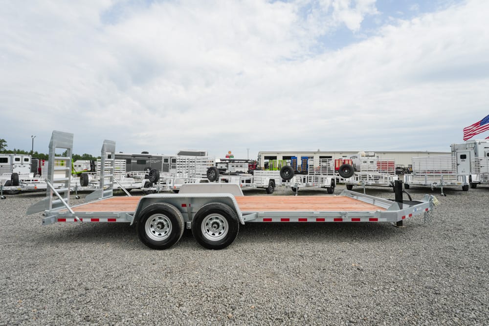 Aluminum car hauler trailer with wooden deck on a gravel lot, rows of similar trailers in the background and an American flag to the right.