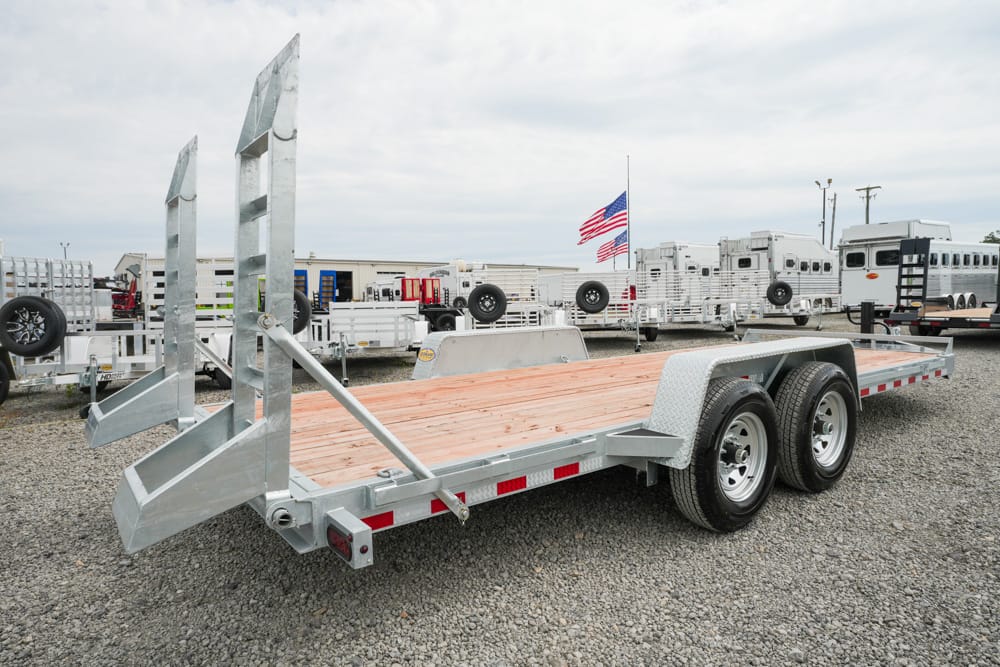 Metal flatbed car trailer with dual axles and wooden deck in a trailer lot; American flag visible in the background