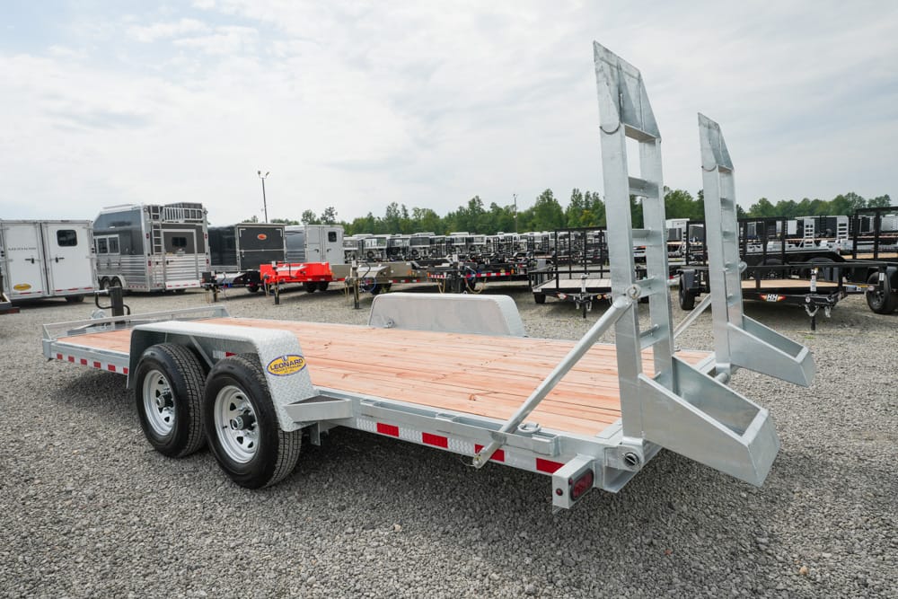 Dual-axle flatbed trailer with a wooden deck and raised rear ramps in a gravel trailer yard