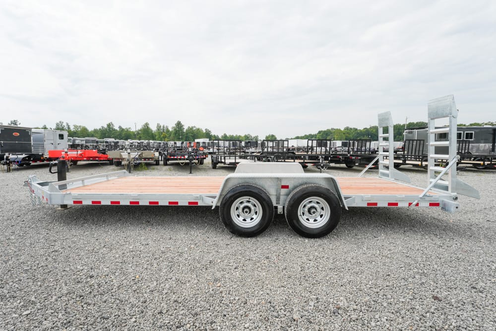 Two-axle flatbed trailer with a wooden deck and raised rear ramps in a gravel trailer yard.