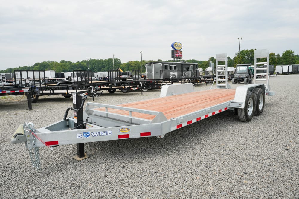 Metal flatbed trailer with a wooden deck and dual axles parked in a gravel lot at a trailer dealership. It has tie-down rails, a front hitch, and a bowise logo on the side.