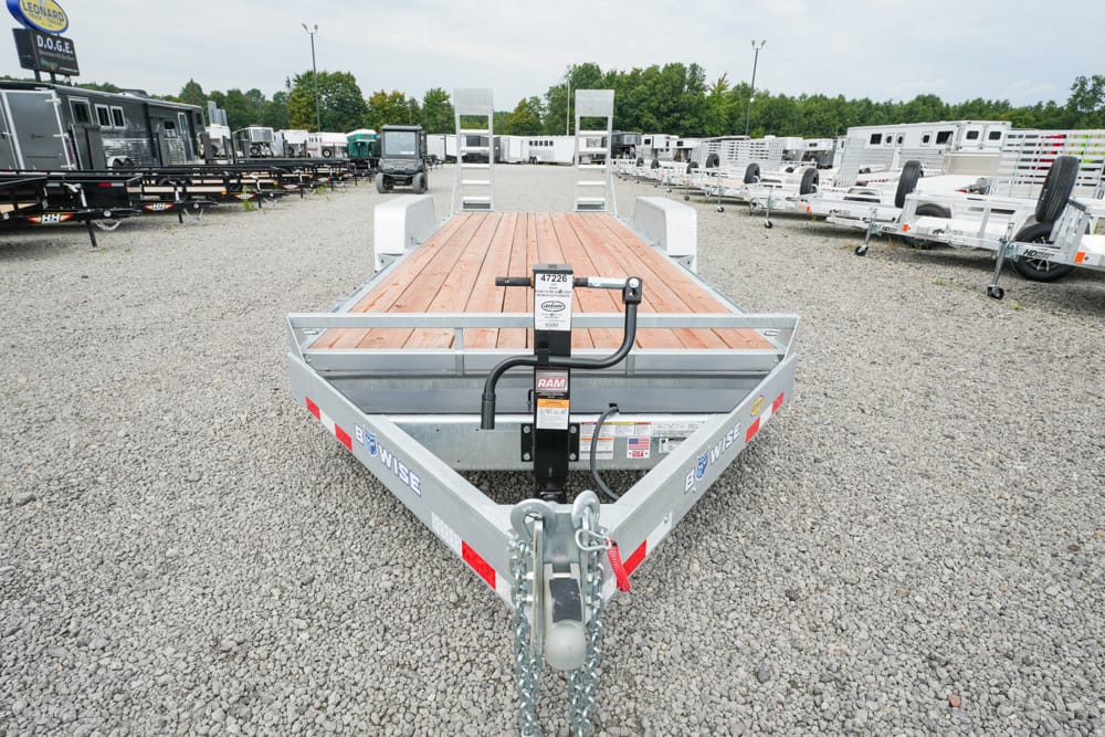Front view of a metal flatbed trailer with a wooden deck in a gravel trailer yard, showing hitch and safety chains on the bow.