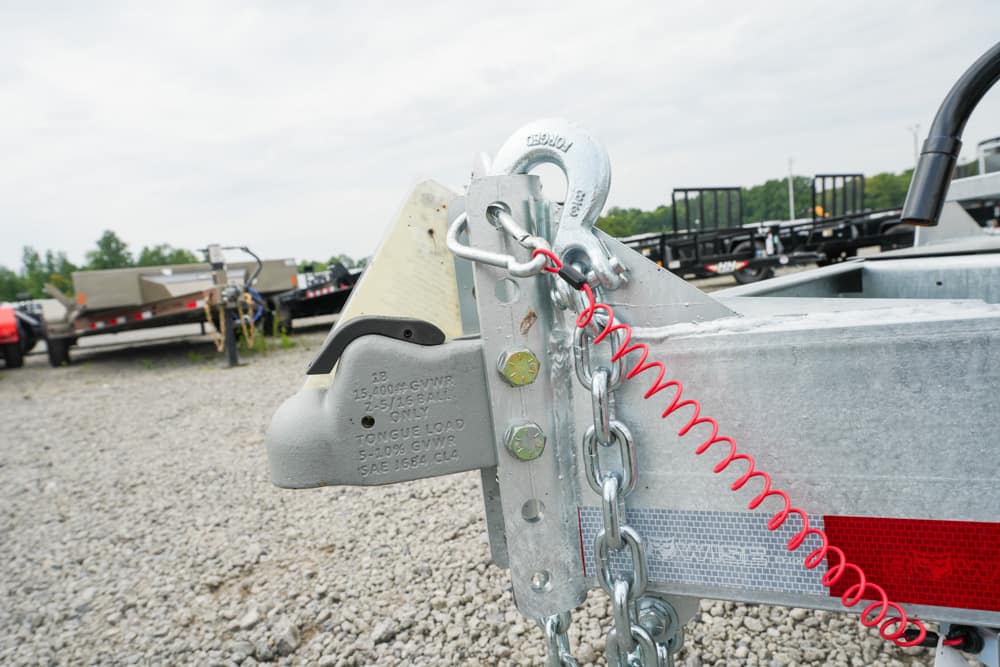 Trailer hitch with safety chain and red coil on a utility trailer in a gravel lot, ready to be connected to a vehicle edge view of other trailers in background (informational image).