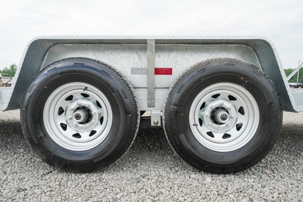 Dual-axle trailer wheels with silver rims on a gravel surface, under a metal fender.