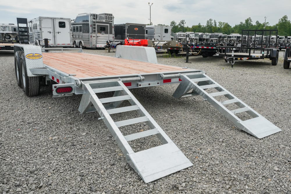 Flatbed trailer with wooden deck and two metal loading ramps in a gravel lot, surrounded by horse trailers and equipment.