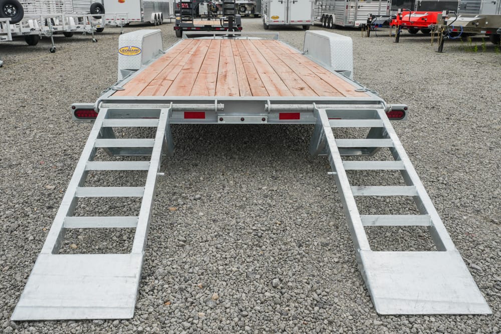 Rear view of a flatbed trailer with a wooden deck and two metal loading ramps extended on a gravel lot in a trailer yard.