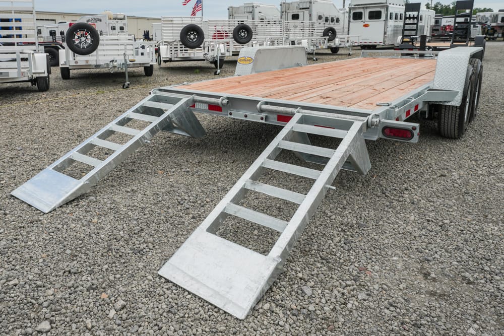 Metal flatbed trailer with two folding ramps extended, wooden deck, on a gravel lot among other trailers.