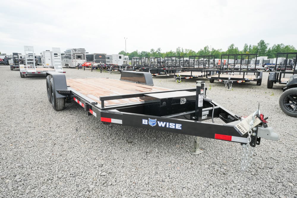 Flatbed utility trailer with a wooden deck on a gravel lot, dual axles, and a front jack stand marked BWise logo on the side.