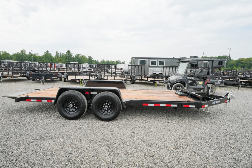 Flatbed trailer with dual rear axles and a wooden deck parked on a gravel lot among other trailers and equipment