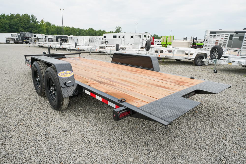 Dual-axle flatbed trailer with a wooden deck and diamond-plate ramps, parked on a gravel lot.