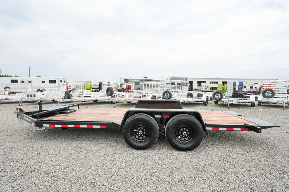 Dual-axle flatbed trailer with a wooden deck on a gravel lot, surrounded by other trailers and equipment in the background.