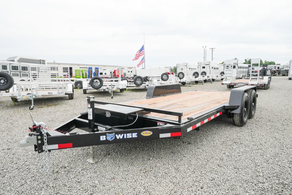 Flatbed trailer with a wooden deck and dual axles sits in a gravel lot among other trailers, American flag in the background.