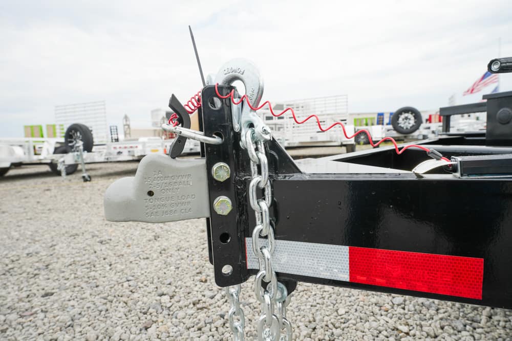 Trailer hitch with a heavy padlock and chained securing hardware on a black trailer frame in a gravel lot with red reflective tape nearby
