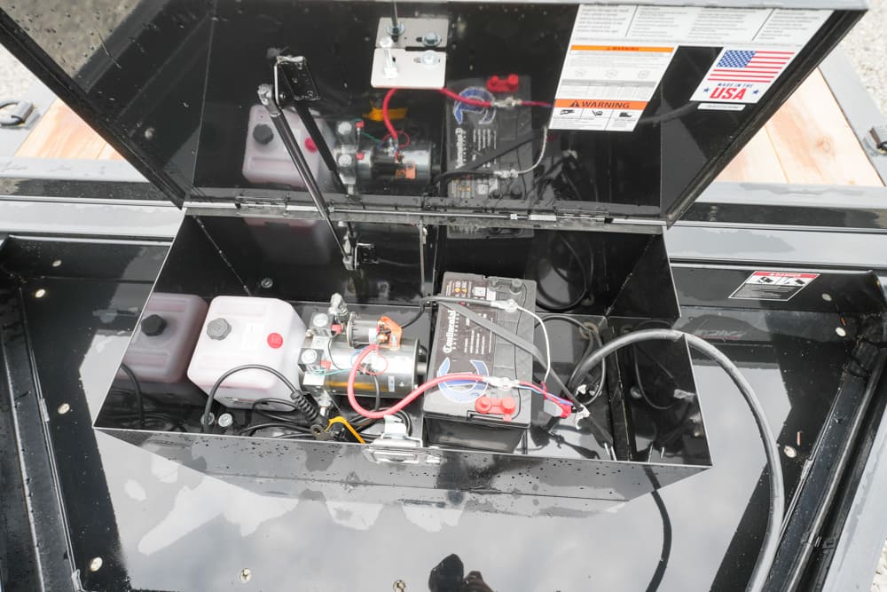 Open compartment showing a small battery box with wired connections, fluid reservoirs, and control components inside a portable generator or power unit with its lid raised.