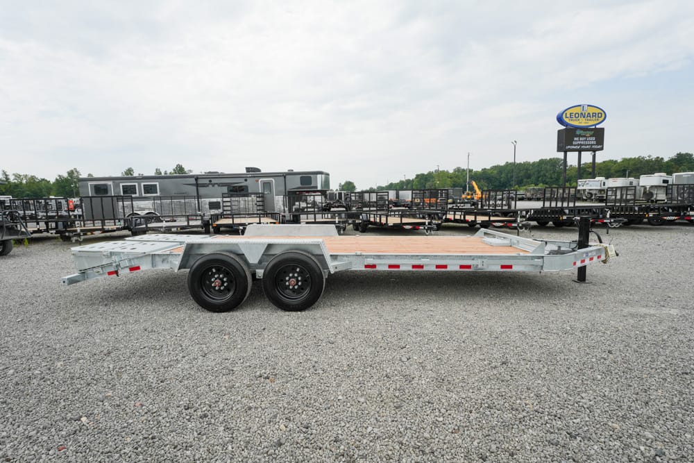 A silver dual-axle flatbed trailer on a gravel lot, with other trailers and a Leonard sign in the background.